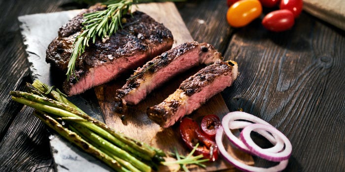 Sliced steak on a wooden cutting board topped with rosemary and accompanied by asparagus and sliced red onion