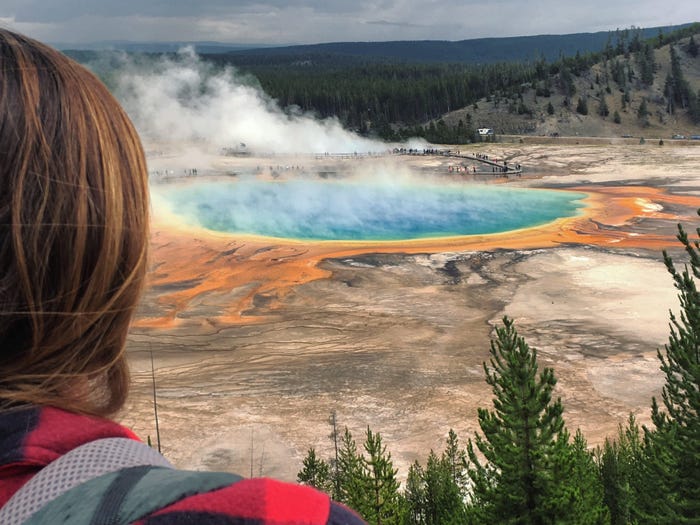 Emily, wearing a black-and-red-checkered jacket and a backpack, looks out at the Grand Prismatic Spring at Yellowstone National Park.