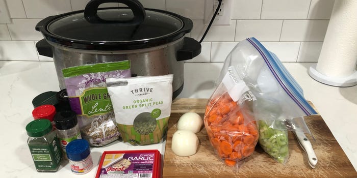 A Crock Pot, seasonings, a container of crushed garlic, bags of lentils and split peas, and a cutting board on a white counter. On the cutting board is an onion sliced in half, a bag of chopped carrots, a bag of celery, and a knife.