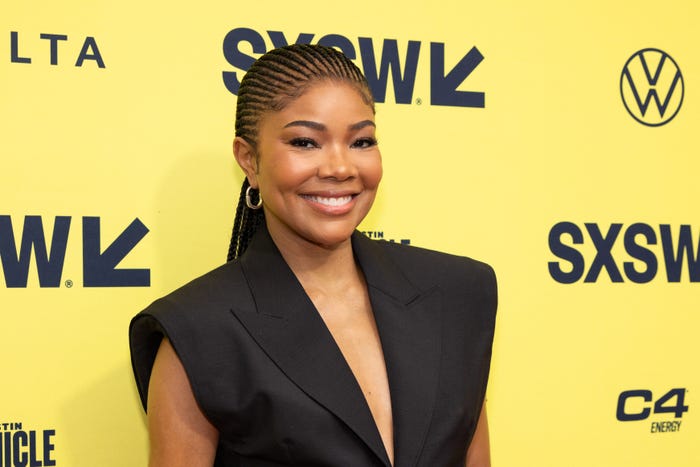 gabrielle union on a south by southwest red carpet, wearing a sleeveless tuxedo shirt and msiling widely with her hair worn in braids pulled back into a ponytail