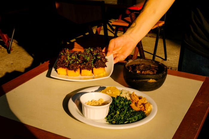 A wood table with a plate with rice and greens, fried polenta with oxtail, and feijoada in a bowl.