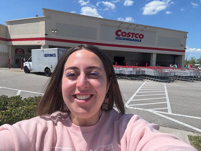 Selfie of the writer in a pink sweatshirt in front of Costco store with bright blue sky in the background