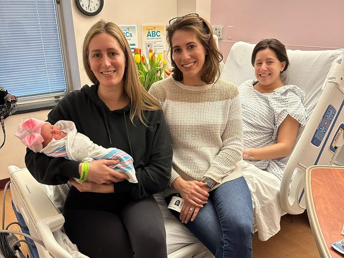 Three sisters are sitting on a hospital bed. One is cradling a baby