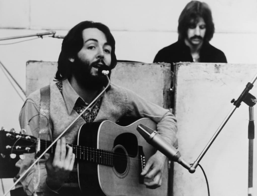 Paul McCartney plays guitar and sings with Ringo Starr in the background of a still from ‘Let it Be’