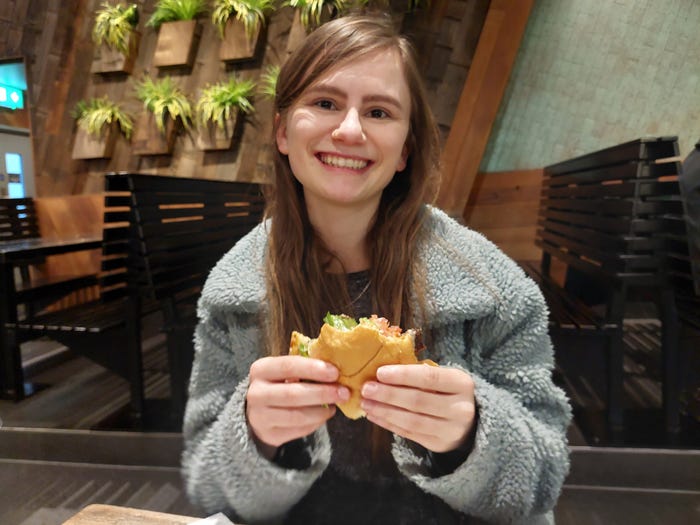 Grace Dean smiling while holding a Shake Shack burger that has been bitten into