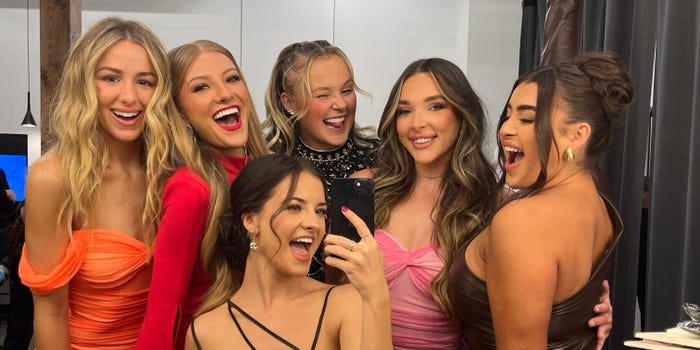 chloe, paige, jojo, kendall, kalani, and brooke from dance moms posing together as young aults. they're all young women wearing dresses, posing together for a selfie in a production studio