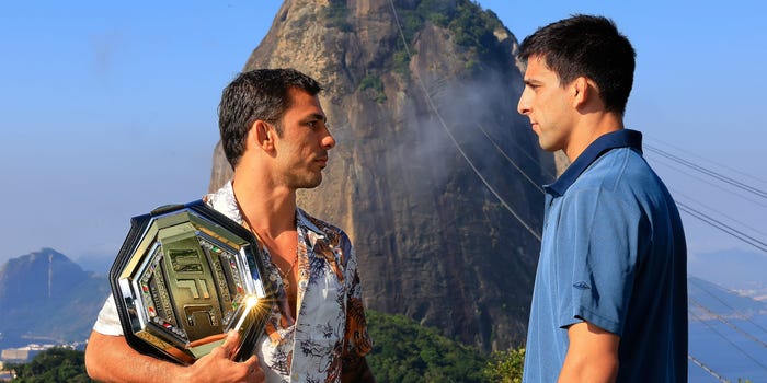 Two men face each other. One man has a UFC belt displayed on his chest.