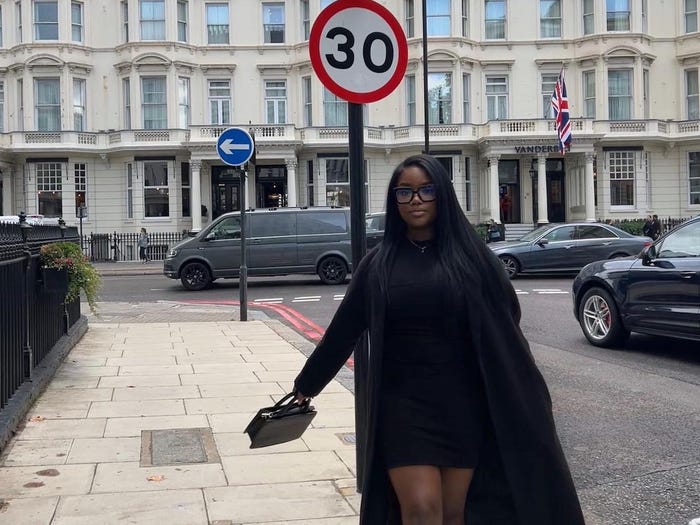 Gladys Nkengasong  wears a black dress and coat and stands on a London street. A building behind her has a a Union Jack flag hanging from it.