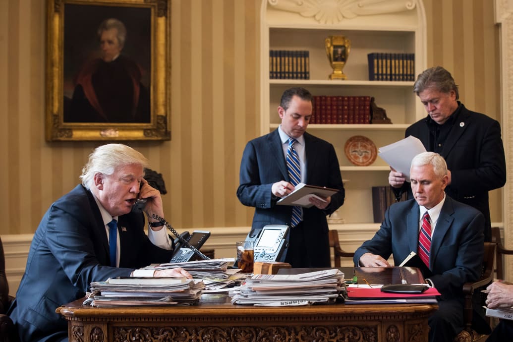 Donald Trump speaks on the phone in the Oval Office. Mike Pence is in a chair facing him and in the background Steve Bannon, dressed all in black, is holding a piece of paper.