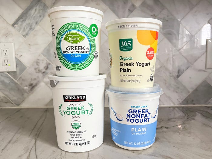 Four containers of yogurt, stacked in two rows, on a gray countertop. They include a white container of Kirkland Signature organic plain Greek yogurt, a white, blue, and green container of Wegmans organic plain nonfat Greek yogurt, a yellow container of 365 organic plain Greek yogurt, and a blue and white container of Trader Joe's plain nonfat Greek yogurt