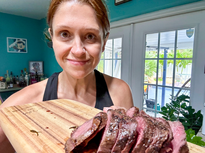 The writer wears a black tank top and smiles while holding a wooden cutting board of rare filet mignon steak slices