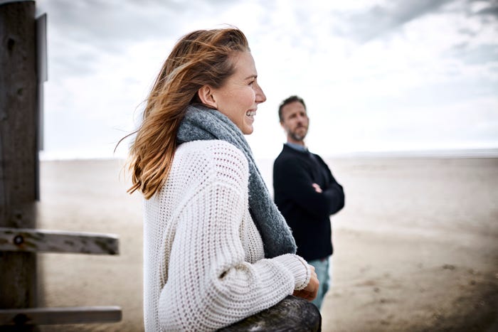 Woman and man wearing sweaters, standing on beach. They are looking at each other and smiling.