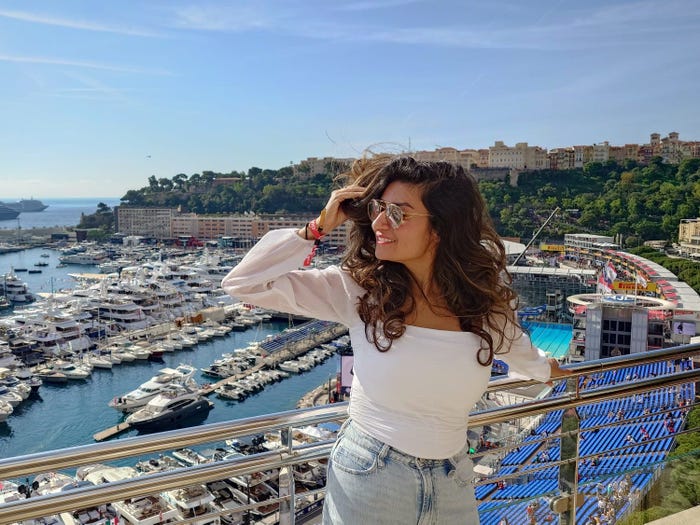 The writer, wearing blue jeans and a white top with her hair blowing in the wind, poses in front of the French Riviera, filled with boats and yachts, with the Grand Prix race track and stands behind her