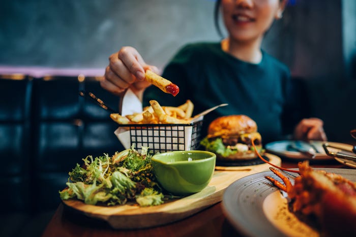 A woman eating a fry from a wooden board with salad and a burger on it.