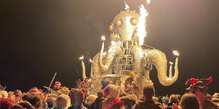 A crowd of dancers at Burning Man festival, surrounding a metal sculpture of an octopus shooting flames mounted on a car