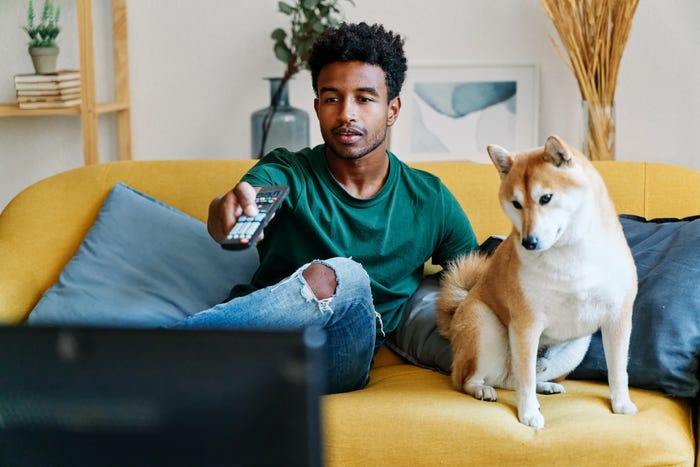 A man, sitting on a couch with a dog, points a remote control at a TV.