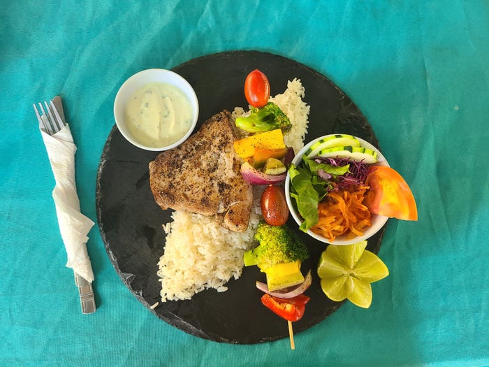 A black plate on a blue tablecloth, featuring a small side salad, white rice, a kebob of vegetables, and a steak of tuna fish.