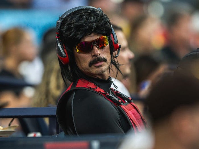 Dr Disrespect wearing a black wig, sunglasses, and mustache sitting at a baseball game and looking directly at the camera,..